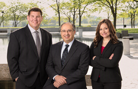 3 people in business attire pose beside a reflecting pool and tree-lined plaza.